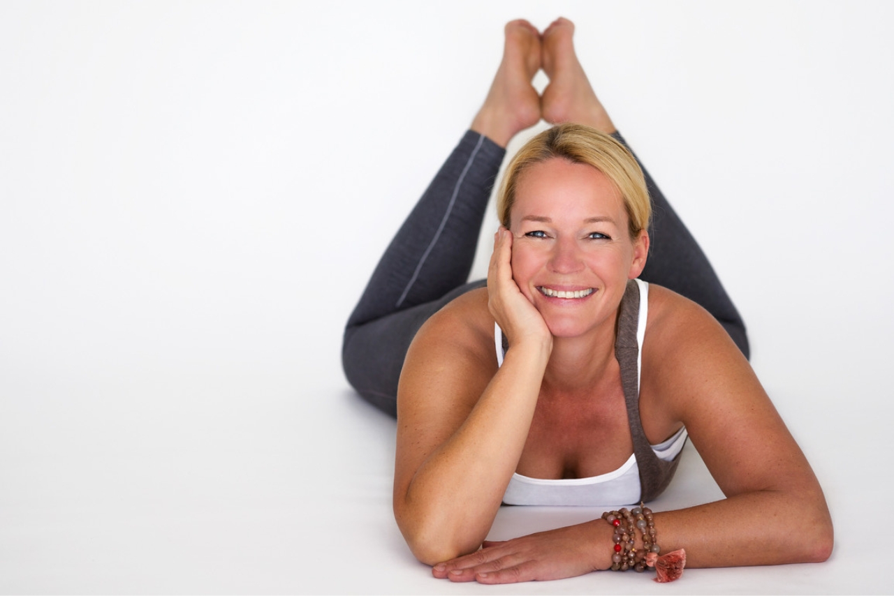 Bettina Bantleon - Yogalehrerin für Hormonyoga in München liegt auf dem Bauch mit angewinkelten Beinen und lächelt in die Kamera.