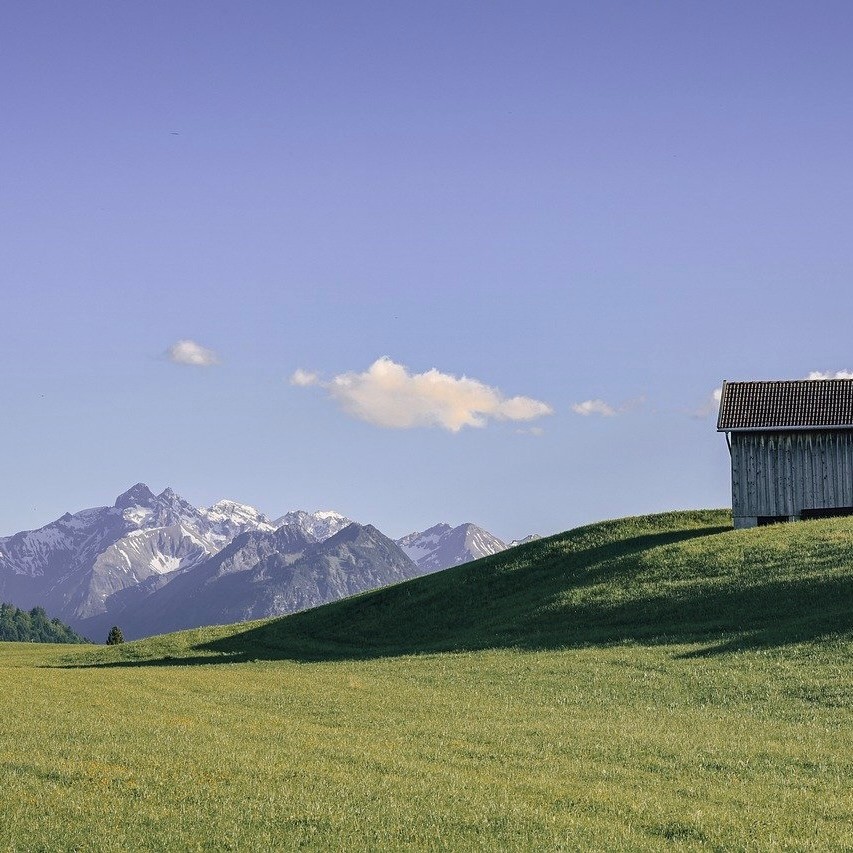 Weite Berglandschaft mit grüner Almwiese, Berghütte und Blick auf alpine Gipfel – ruhige Naturkulisse für ein exklusives Yoga-Retreat in den Bergen, ideal für Entspannung, Achtsamkeit und nachhaltige Erholung.